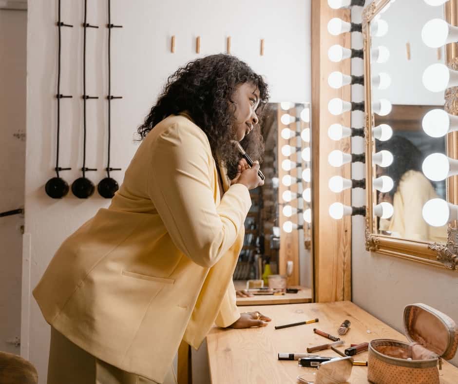 A lady applying make up in front of a rectangular mirror while holding onto a dresser