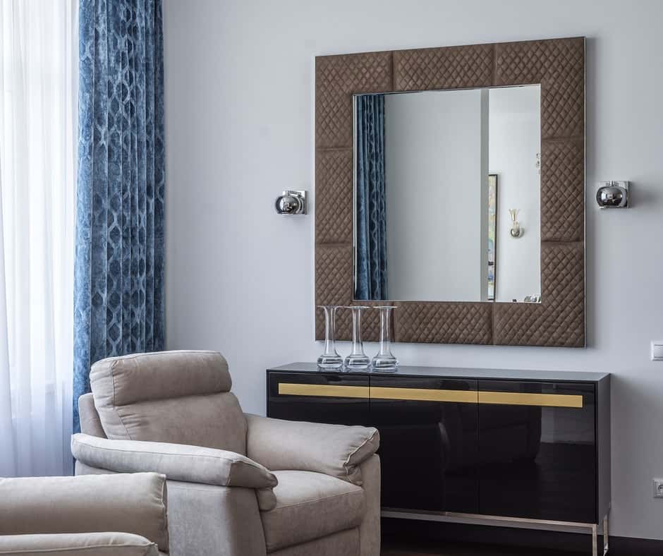 A room with a chair and dresser with a square mirror hang on top of it.